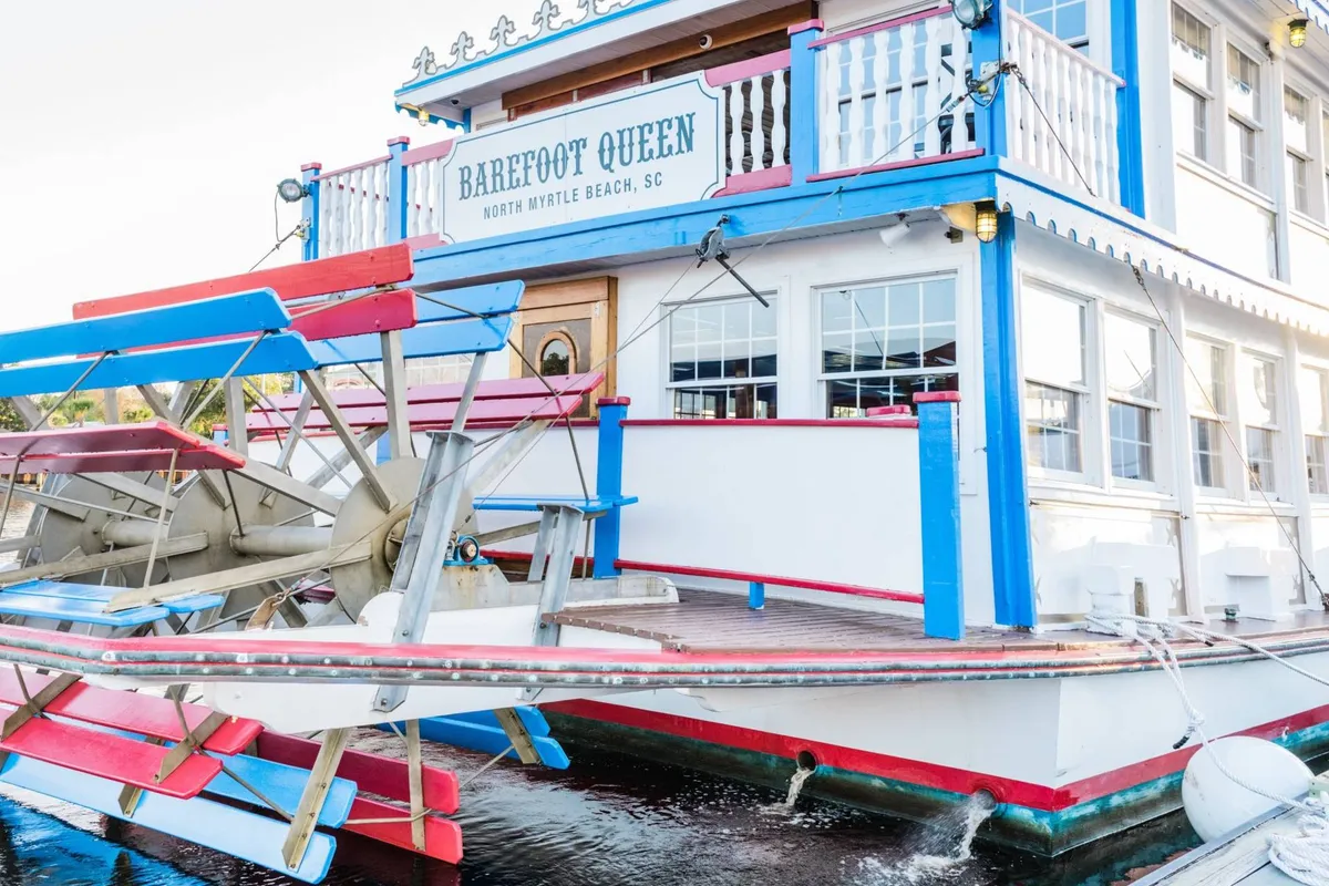 The colorful exterior of the Barefoot Queen Riverboat, prominently featuring its paddle wheel and inviting facade. The image captures the festive atmosphere and waterfront setting, hinting at the scenic cruises and family gatherings that await.