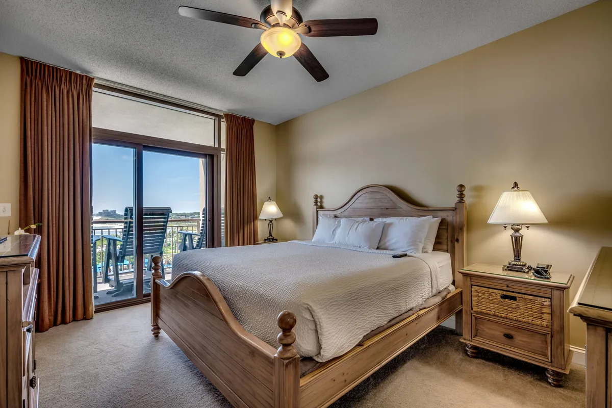 A serene bedroom featuring a large, inviting bed with plush linens, two nightstands with lamps, and a private balcony offering a gentle view. The calming color palette and soft lighting evoke comfort and relaxation, while the TV and balcony doors hint at privacy and a connection to the outdoors.