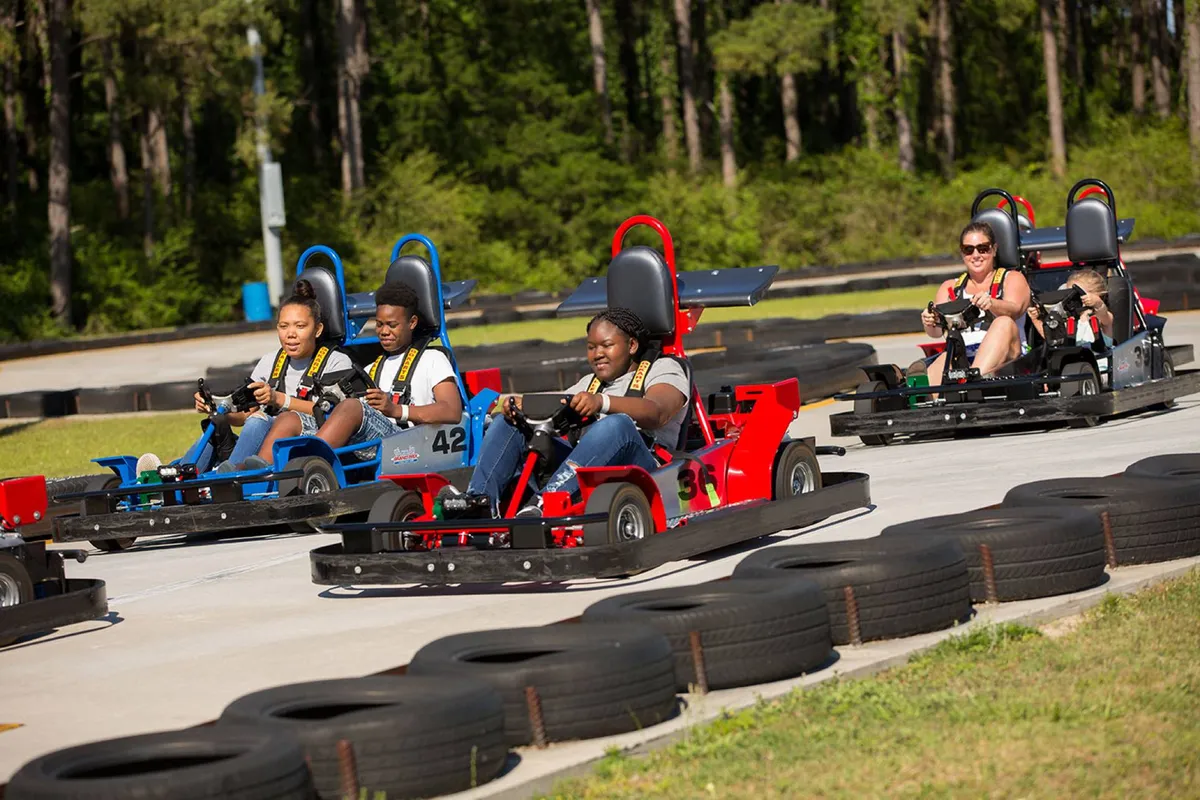 A dynamic outdoor go-kart racetrack with families and groups racing, surrounded by safety barriers. The scene is active and playful, with smiles and excitement as participants compete and enjoy the friendly competition.
