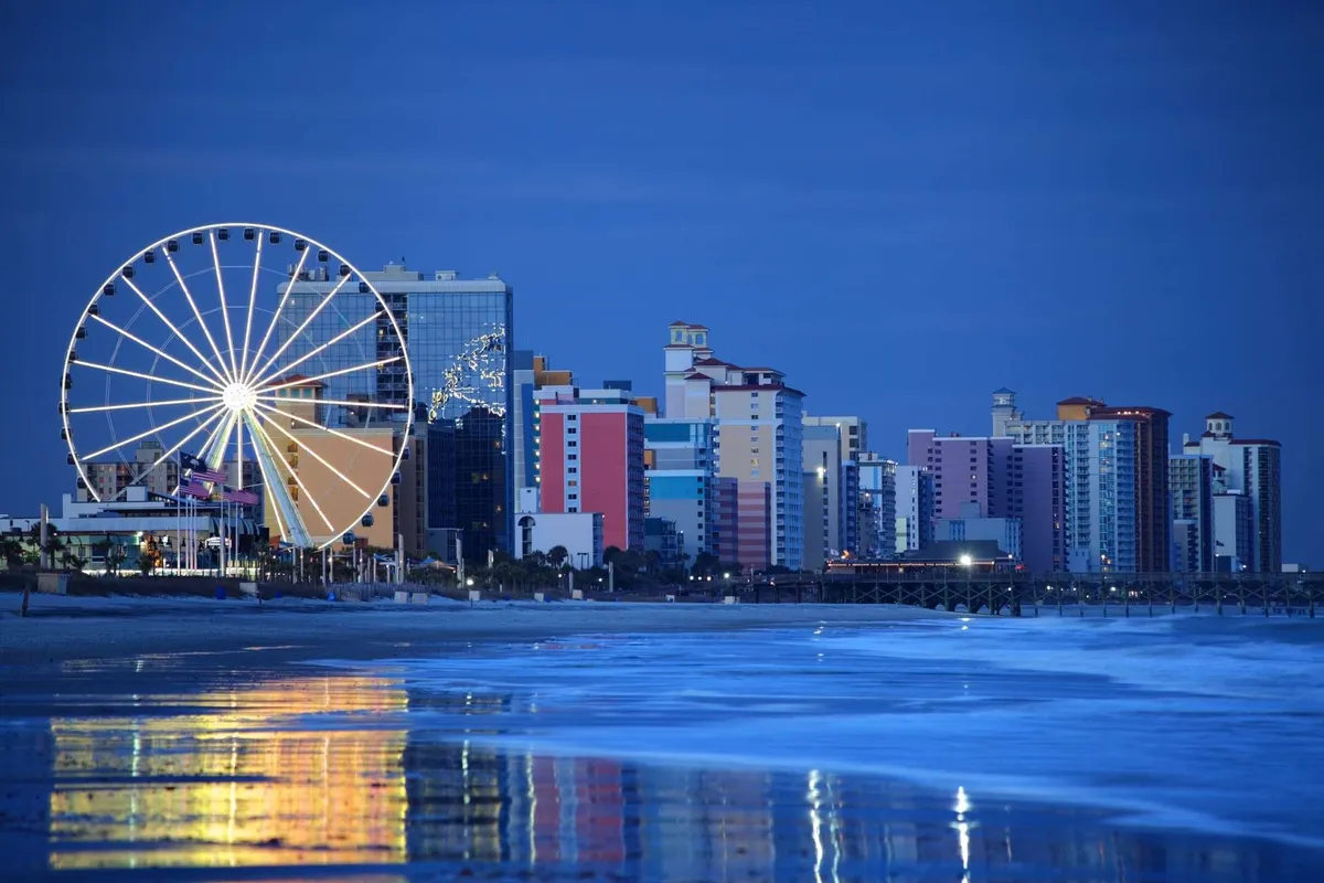A lively waterfront view featuring a colorful Ferris wheel, vibrant buildings, and reflections in the water. The scene is festive and bustling, representing the nearby entertainment and dining options at destinations like Barefoot Landing, which are ideal for families seeking shared experiences and fun for every generation.