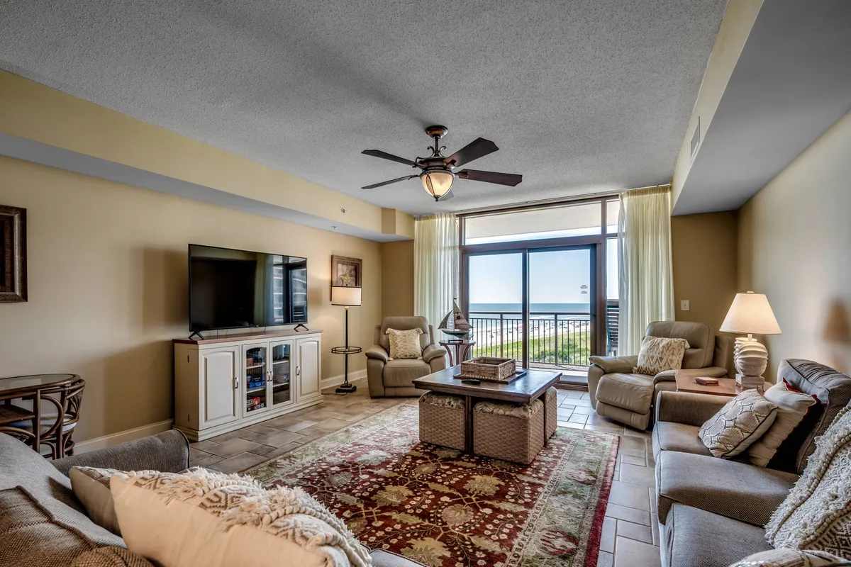 This image showcases a spacious, sunlit living area with plush, comfortable seating arranged around a sleek coffee table and a modern TV unit. Floor-to-ceiling windows reveal stunning sunset views, filling the space with a warm, inviting glow. The setting exudes coziness and is perfect for relaxing, socializing, or simply soaking in the beautiful North Myrtle Beach scenery.