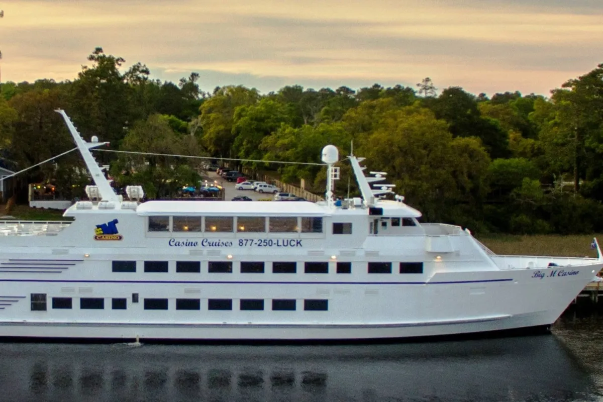 A large white cruise ship is docked on calm waters, surrounded by lush trees. The deck is set for socializing, dining, and enjoying scenic sunsets, with a sense of relaxation and upscale leisure throughout the scene.