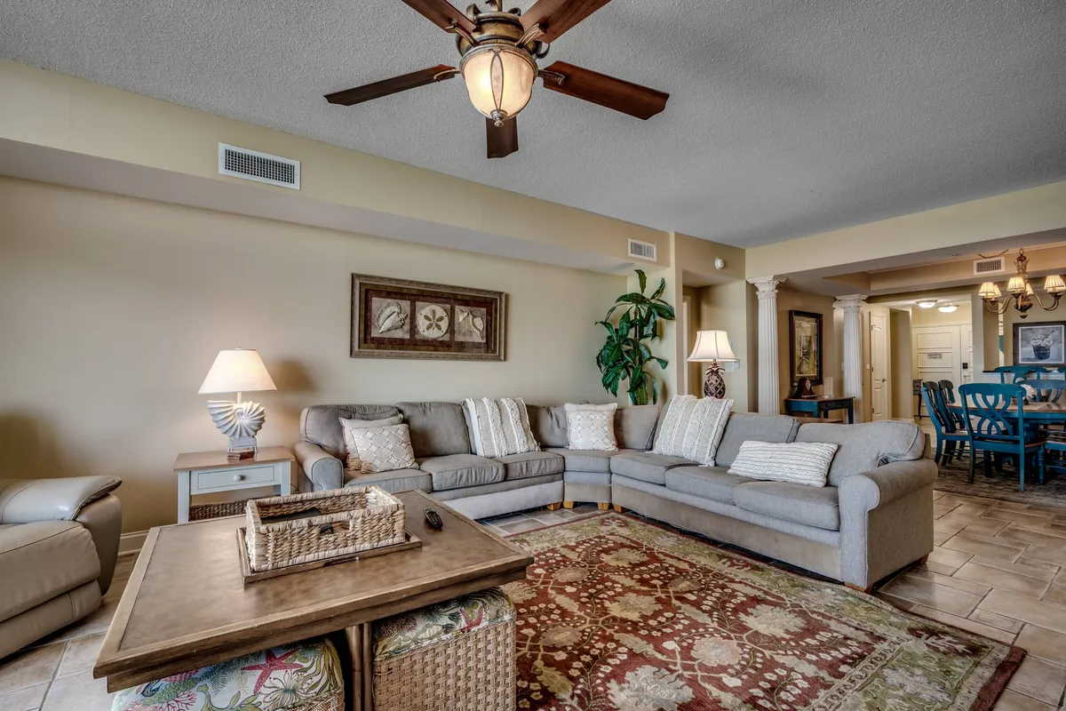 A spacious living room featuring a large sectional sofa, decorative coffee table, ceiling fan with a light fixture, framed wall art, and an indoor plant. The arrangement is open and inviting, ideal for gathering and relaxing with family and friends.