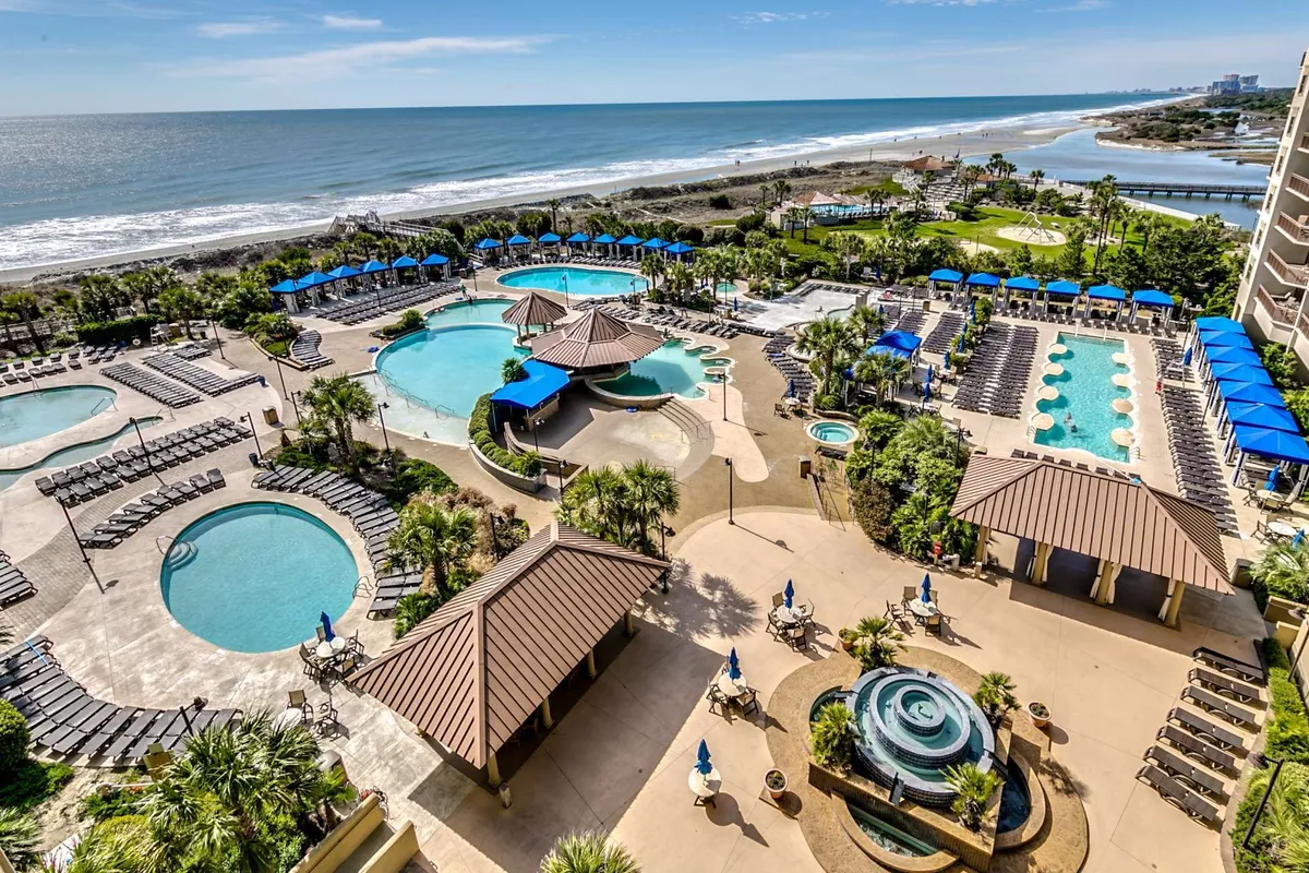 A sweeping view of the resort's main pool complex, featuring multiple swimming pools, landscaped gardens, lounging areas, and direct beachfront access. The vibrant scene includes families and groups enjoying the water, sunbathing, and relaxing by the pools amidst lush greenery and elegant water features.