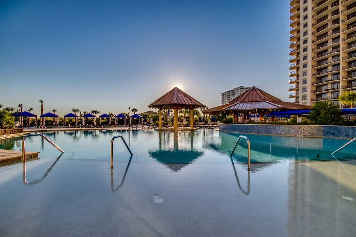 A sprawling outdoor pool area featuring a large swimming pool surrounded by thatched-roof cabanas and plenty of lounge chairs. The sun is setting, casting a warm glow over the scene, with families and guests relaxing poolside, socializing, and enjoying the serene atmosphere.