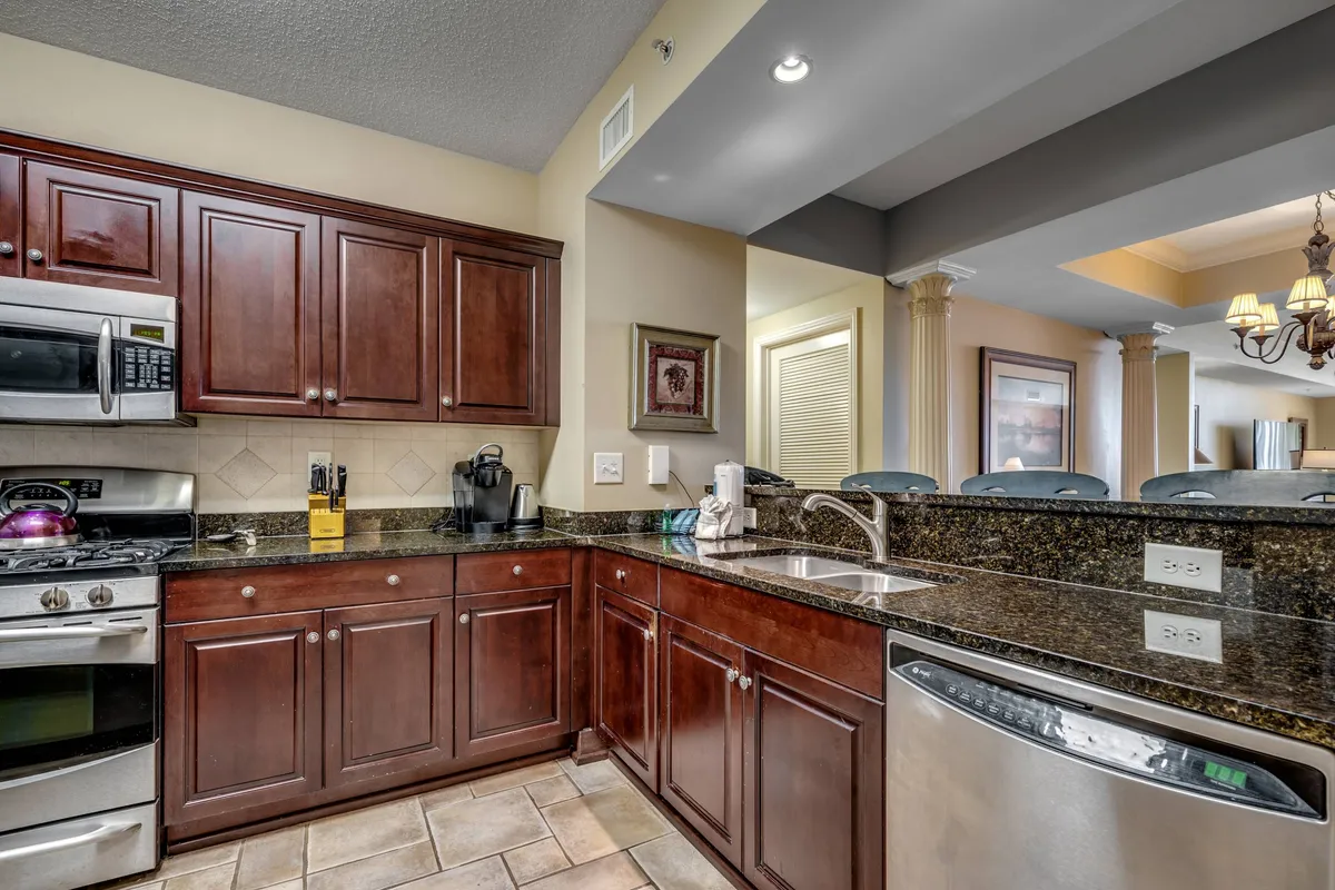 A view of the kitchen showcasing dark wood cabinetry, granite countertops, stainless steel appliances, and a large kitchen island with bar seating. The image suggests an open, sociable layout, ideal for entertaining and casual dining. The space feels warm and inviting, encouraging group cooking and conversation.
