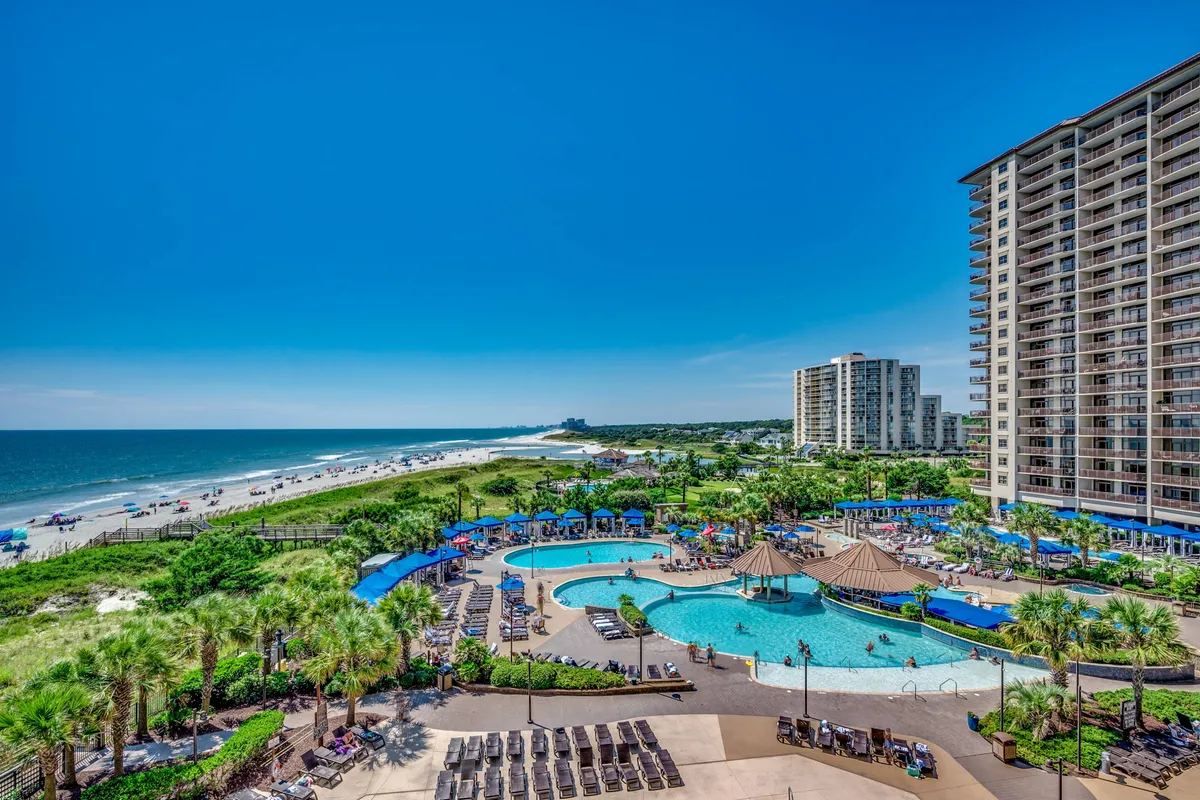 A lively scene of a swimming pool adjacent to the beach and ocean, surrounded by palm trees and nearby resort buildings. The area is animated with guests swimming, sunbathing, and gathering with friends or family, with sunset hues enhancing the cheerful mood.